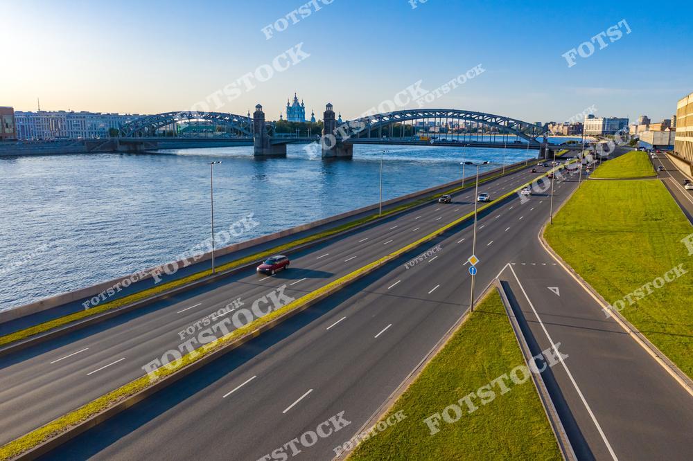 Фотография солнечного летнего дня в Санкт-Петербург, Россия. На изображении представлен вид на реки и мосты Санкт-Петербурга, где в кадре запечатлён Большеохтинский мост, также известный как мост Петра Великого. В пролёте моста хорошо виден силуэт Смольный собор, что создаёт выразительную архитектурную композицию. Фотоснимок передаёт атмосферу ясной летней погоды, сочетание водного пространства, инженерной архитектуры и исторических достопримечательностей города. Это изображение подчёркивает уникальный облик Санкт-Петербурга как города рек и мостов. Иллюстрация может использоваться как картинка или слайд, посвящённые летнему Петербургу и городским пейзажам России.